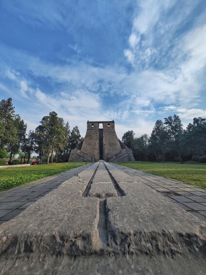 A stone pathway leading to a large, ancient-looking structure with a central opening, surrounded by trees and a clear sky. 3