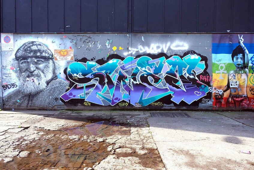 A wall with graffiti, a mural, and a puddle on the ground. 1