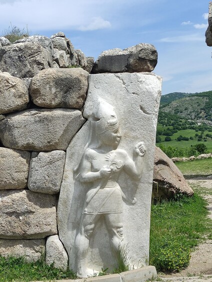 A stone relief of a figure holding a staff, set against a stone wall with a rural landscape in the background. 5