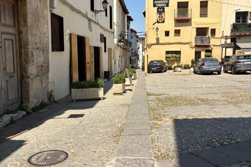 A cobblestone street with buildings, a manhole cover, and parked cars. 1