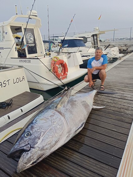 A man kneeling next to a large fish on a dock with boats in the background. 3