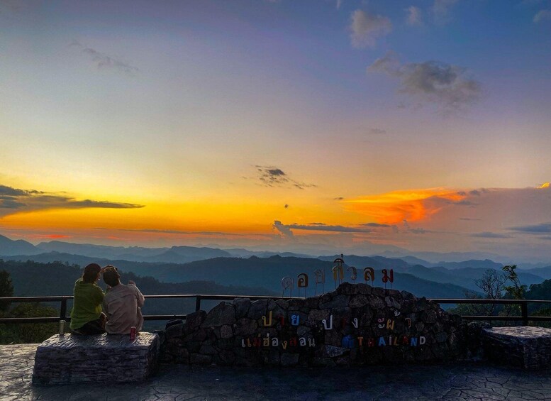 Two people sitting on a stone bench overlooking a mountainous landscape with a sunset. 1