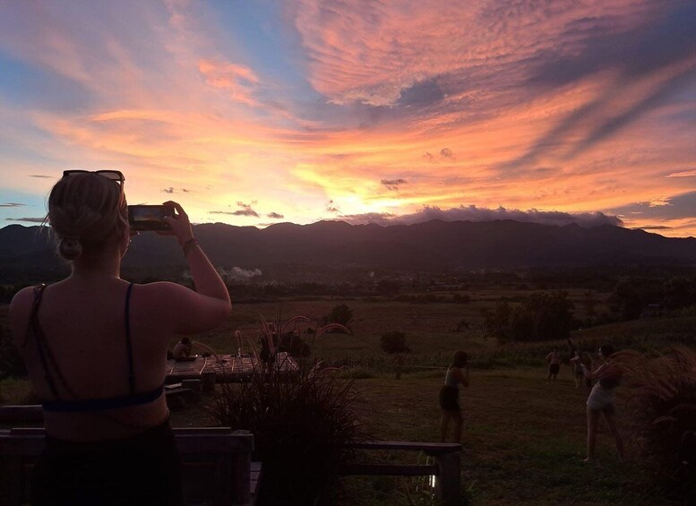 A sunset with a woman taking a photo, mountains in the background, and a grassy field with people. 2
