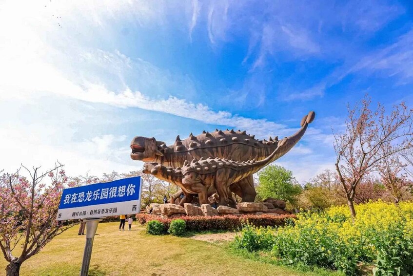 A large dinosaur sculpture in a park with a blue sign and blooming trees. 5