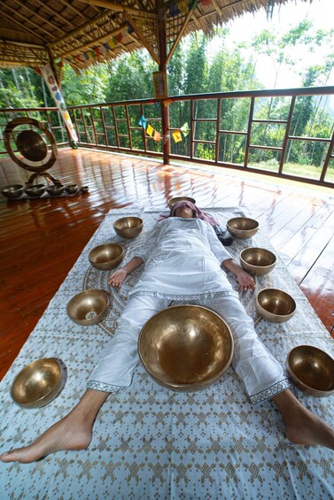 A person lying on a mat surrounded by large bowls, in a room with a wooden floor and a thatched roof. 1
