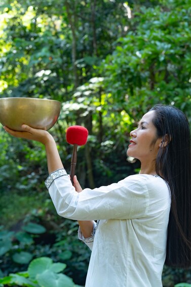 A person holding a large brass bowl and a red mallet, standing in a lush green forest. 3
