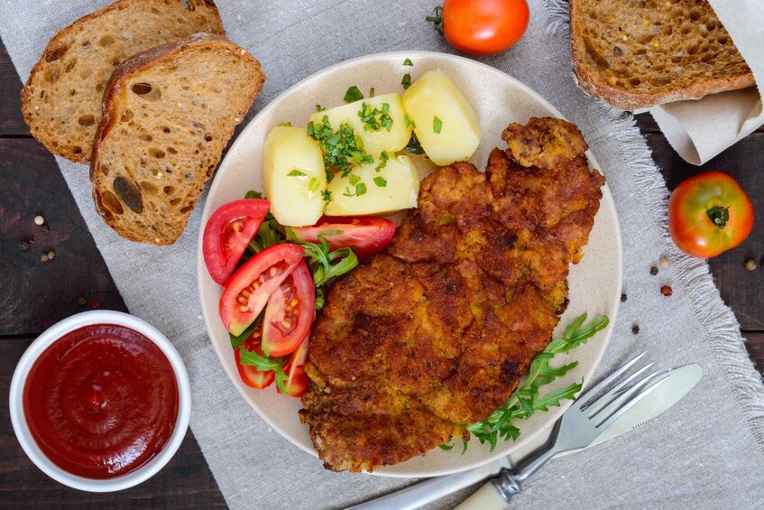 A plate with a breaded cutlet, boiled potatoes, and a salad, accompanied by slices of bread and a bowl of ketchup. 2
