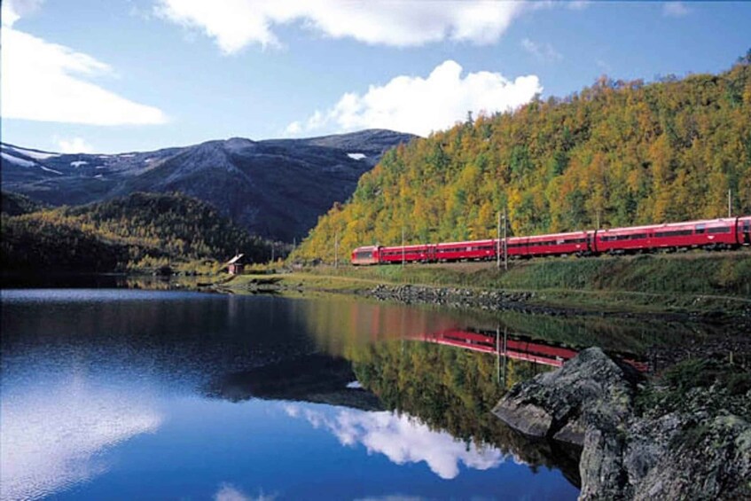 A red train traveling along a track beside a calm lake with autumn-colored trees and mountains in the background. 2
