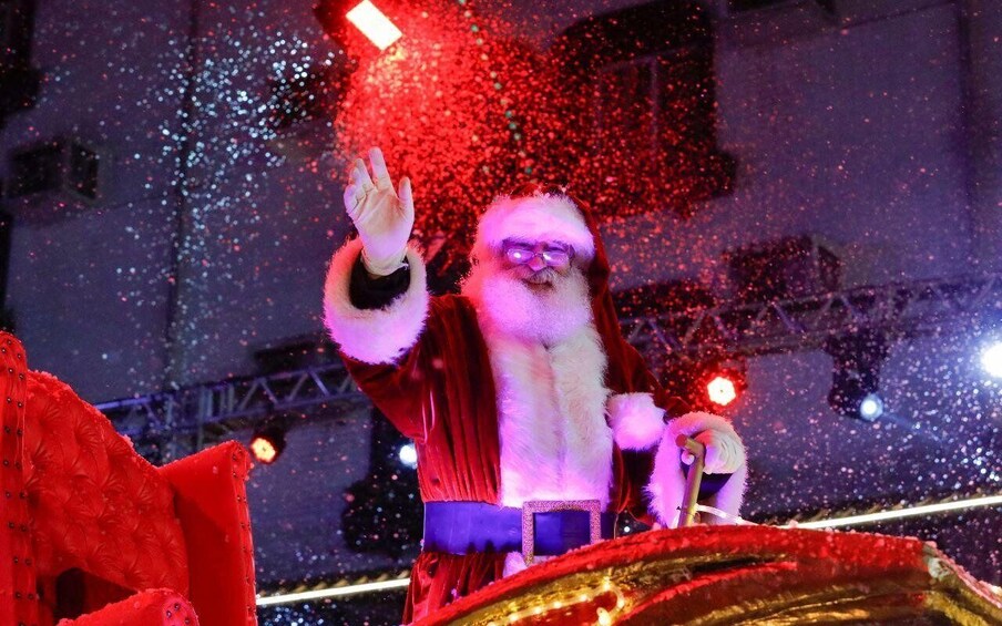 A person in a Santa costume waving, with a red and gold sleigh in the foreground. 1