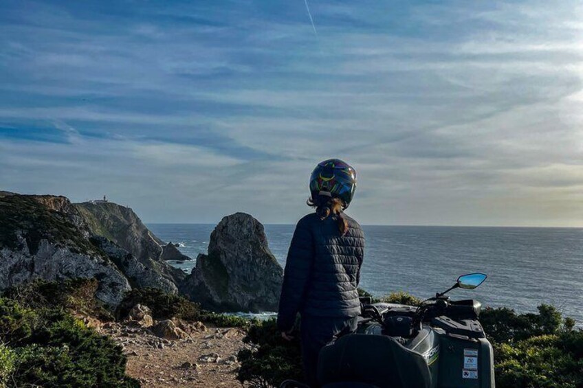 A person with a helmet and backpack standing by a scooter on a rocky coastal path with the ocean in the background. 5