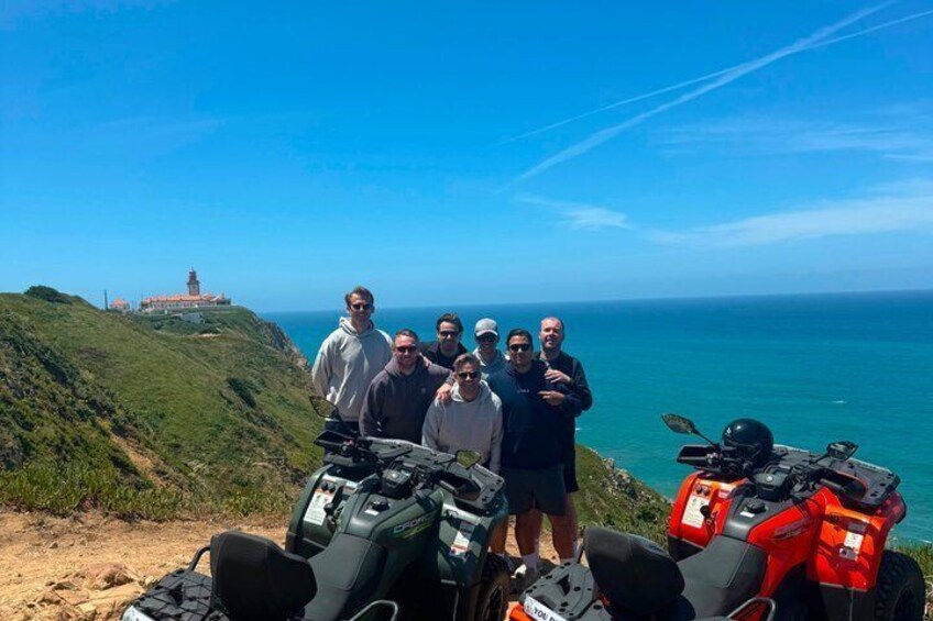 A group of people standing on a cliff with ATVs in the foreground and a lighthouse in the background. 2