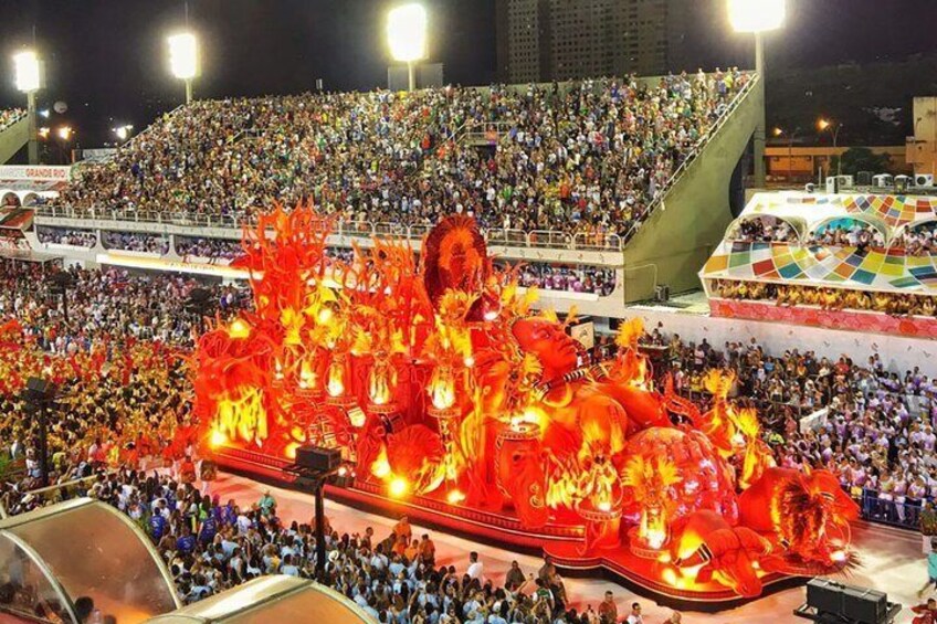 A large outdoor event with a brightly lit float featuring a dragon design, surrounded by a crowd in an open-air stadium. 2