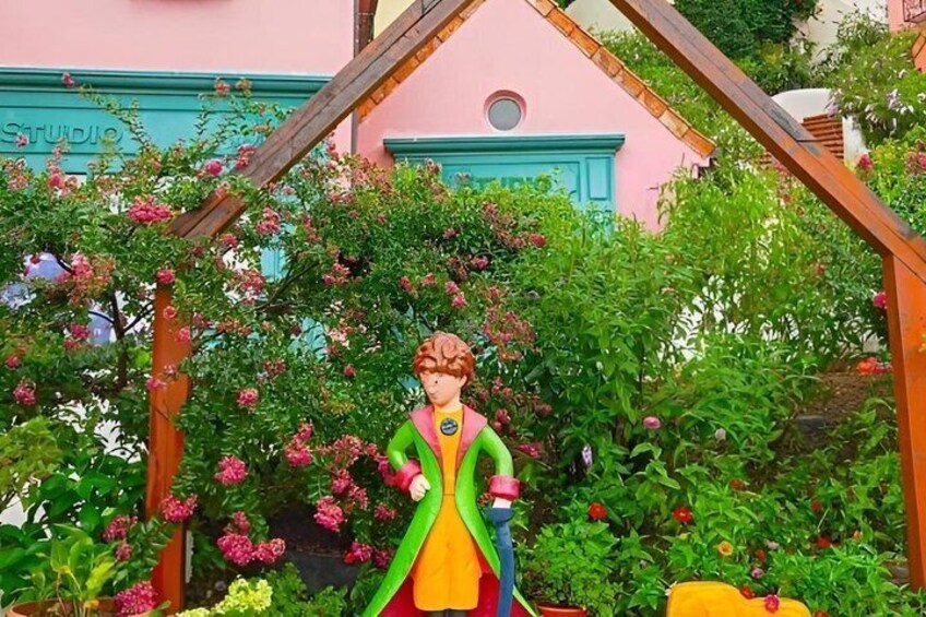 A colorful statue of a woman in a green and yellow dress, surrounded by lush greenery and pink flowers, with a pink building in the background. 2