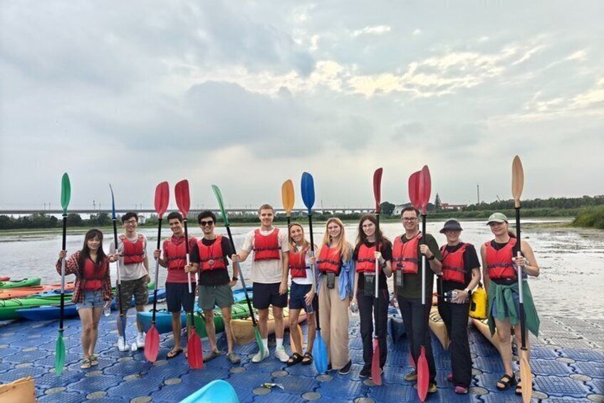 A group of people in life jackets on a boat, holding paddles, with a river and bridge in the background. 2
