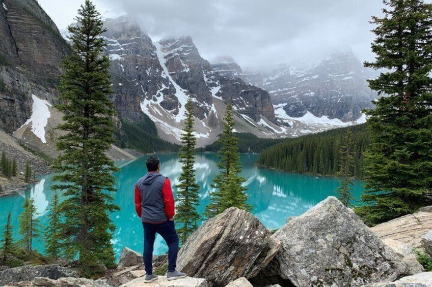 A person standing on a rocky outcrop overlooking a turquoise lake surrounded by mountains and coniferous trees. 1