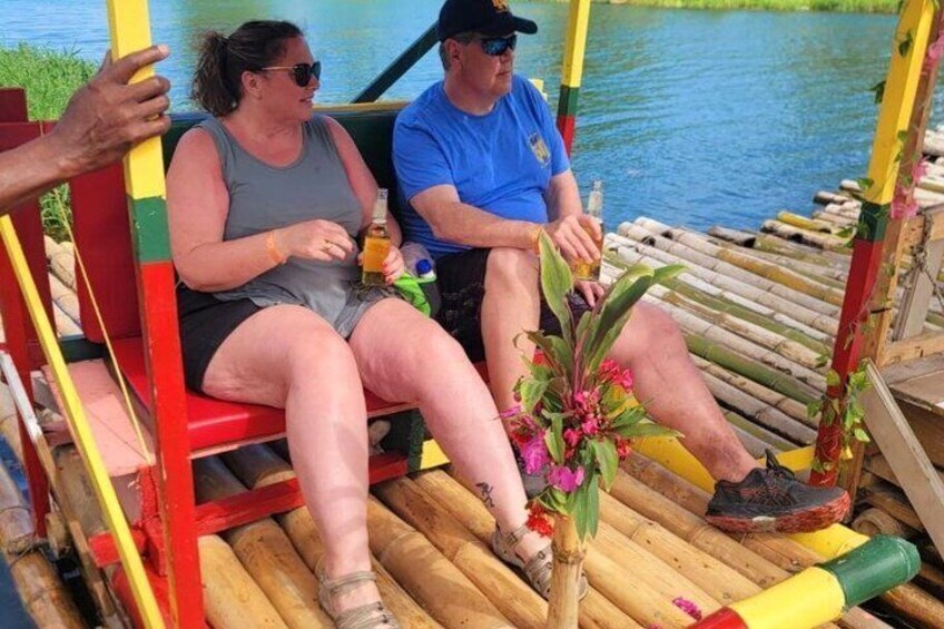 Two people on a wooden boat with a red and yellow structure, holding drinks, and a plant with pink flowers. 4