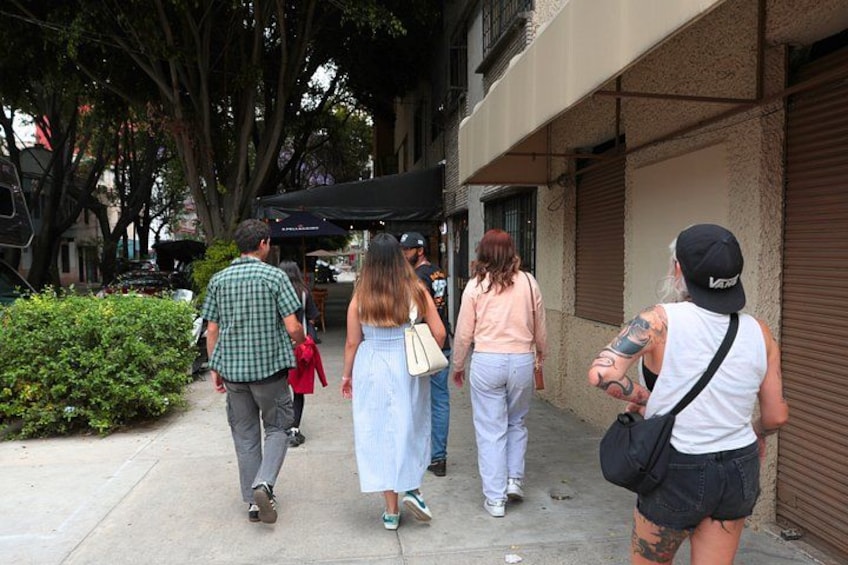 A sidewalk with pedestrians, a building with closed shutters, and a tree-lined street. 5