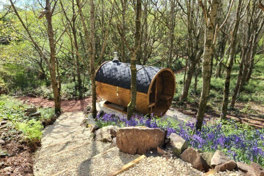 A wooden barrel structure in a forest with a gravel path and purple flowers. 4