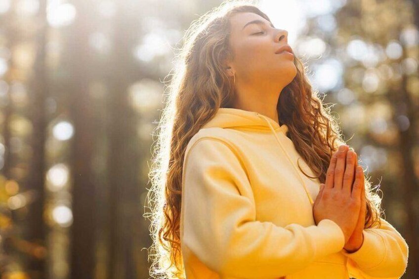 A person in a yellow hoodie with hands in a prayer position, standing in a forest with sunlight filtering through the trees. 1