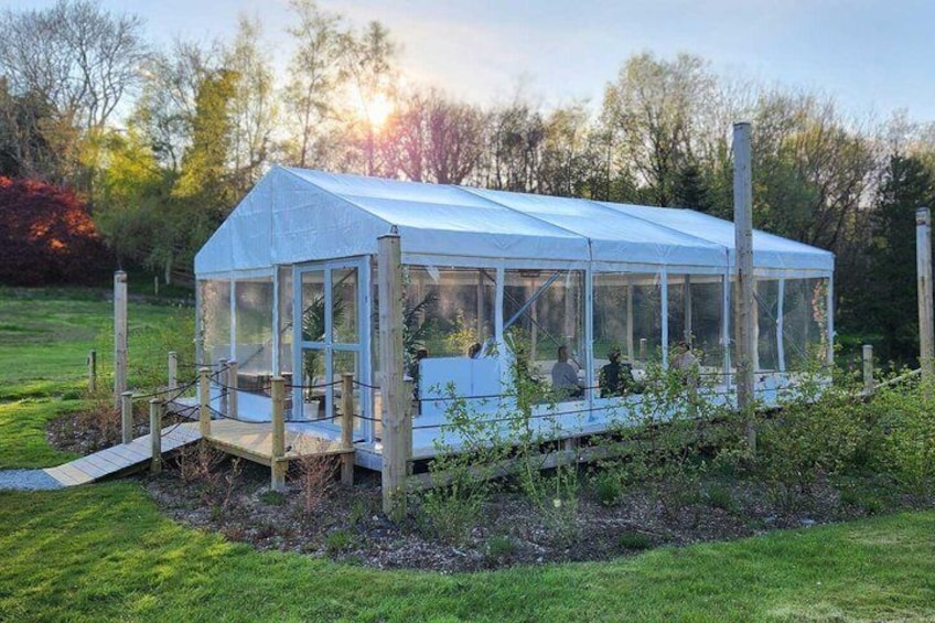A greenhouse structure with a transparent roof, surrounded by a wooden fence, set in a grassy field with trees in the background. 2
