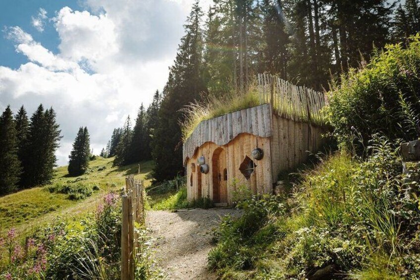 A rustic wooden house with a thatched roof, surrounded by lush greenery and a forested hillside. 5