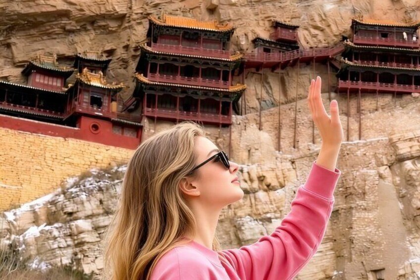 A person in a pink top with a hand raised, standing in front of a cliff with traditional multi-tiered buildings. 2