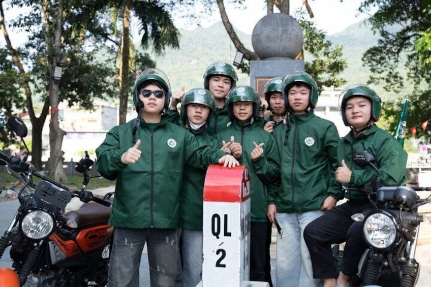 A group of people in green jackets posing with a mailbox and motorcycles. 1