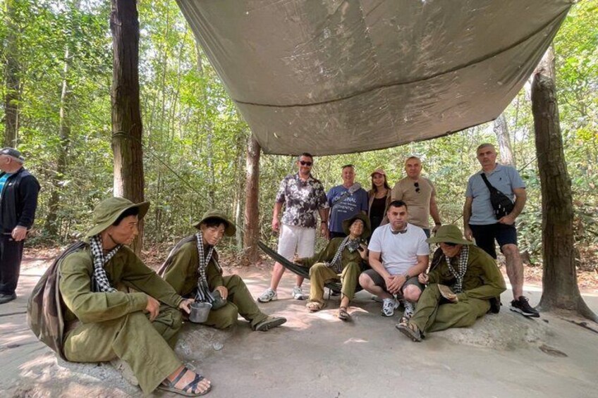 A group of people sitting and standing under a canopy in a forested area. 3