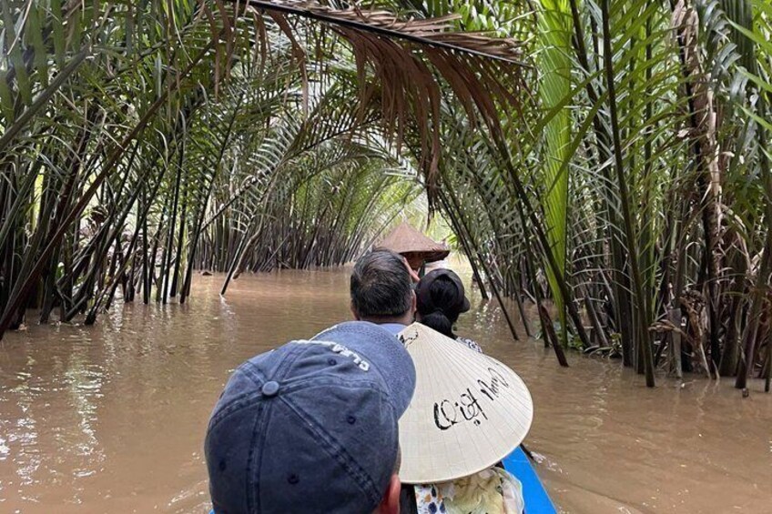 A person in a blue shirt and a straw hat, paddling a boat through a flooded area with tall palm trees. 1