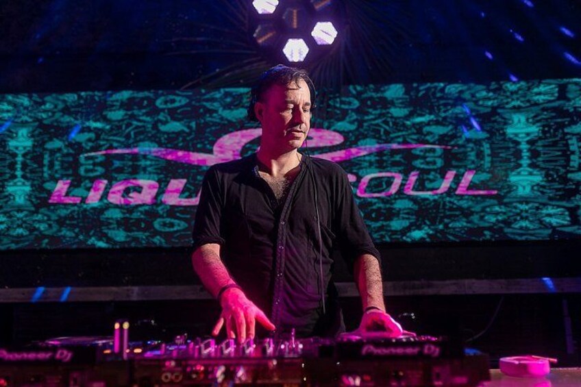 A person standing at a DJ booth with a large screen displaying the word 'LIAO' in the background. 3