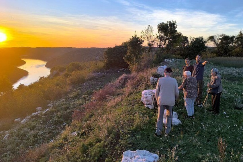 A group of people standing on a grassy hillside overlooking a river at sunset. 5