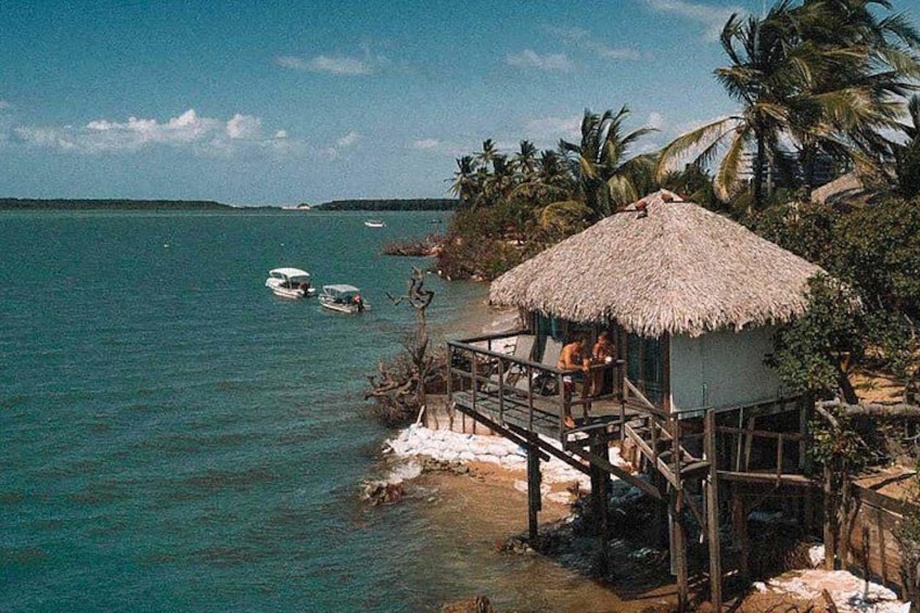 A thatched-roof hut on stilts by a turquoise sea, with boats in the water and palm trees in the background. 2