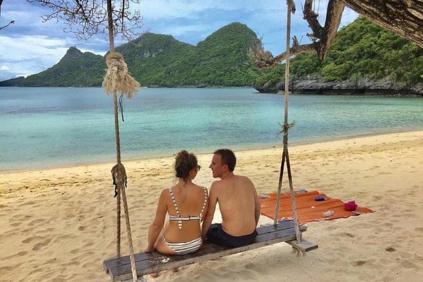 A couple sitting on a wooden swing on a sandy beach with a clear view of the ocean and green hills in the background. 3