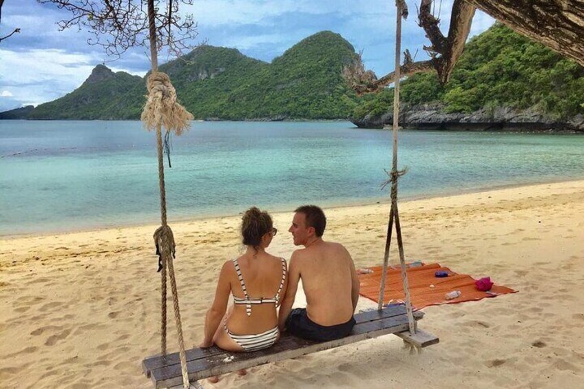 A couple on a wooden swing overlooking a beach with clear water and green hills in the background. 2