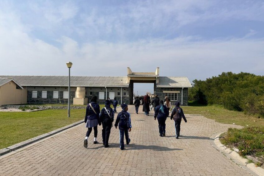 A paved pathway leading to a building with a traditional roof, surrounded by grass and trees. 2