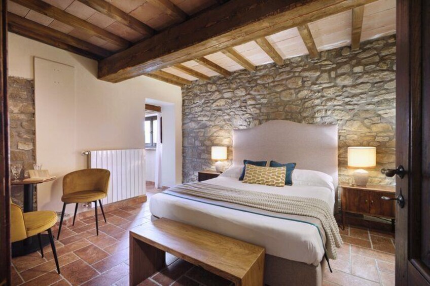 A bedroom with a stone wall, a wooden bed, a bench, and a chair. 5