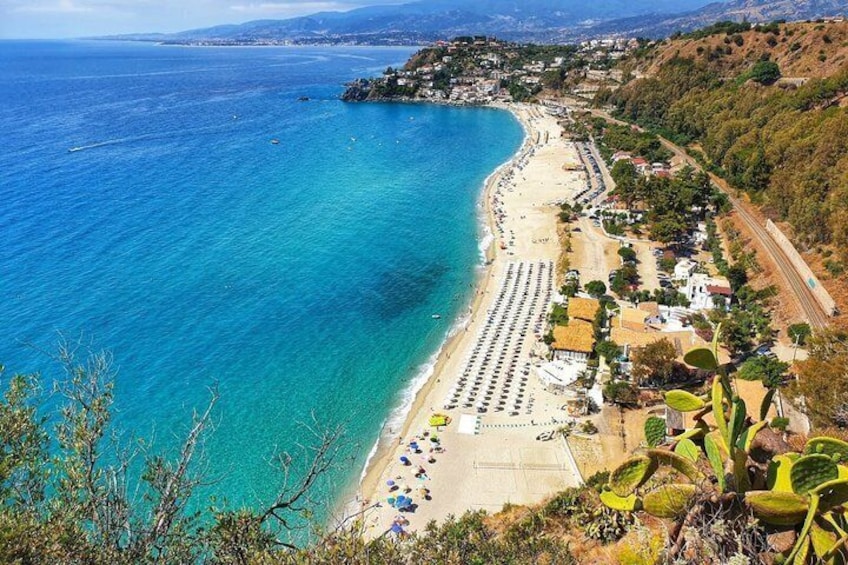 A coastal landscape with a beach, clear blue waters, and a hilly terrain with vegetation. 1