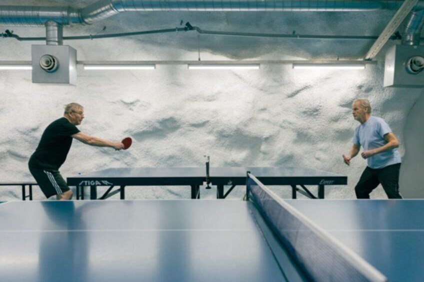 Two people playing table tennis in an indoor setting with a white wall and a table tennis table. 5