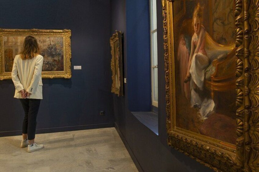 A person observing paintings in an art gallery with dark walls and ornate frames. 5