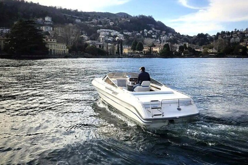 A white motorboat on calm water with a person steering, surrounded by a hilly landscape with buildings. 1