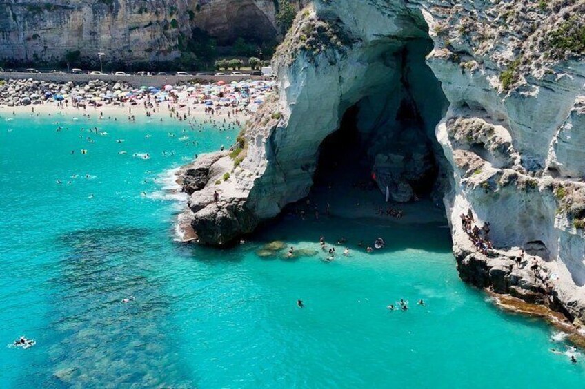 A coastal beach with clear turquoise water, a rocky arch formation, and a crowd of people. 3