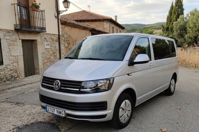 A silver Volkswagen van parked on a rural road with stone buildings and a clear sky in the background. 3