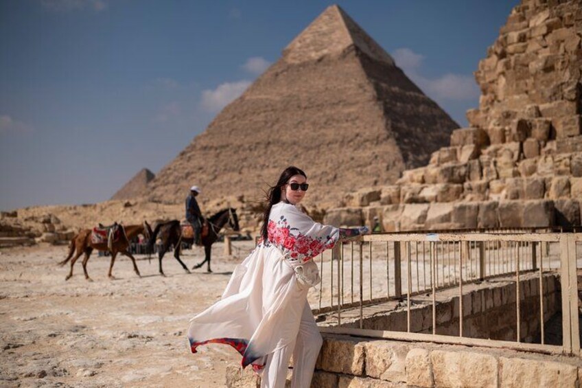Eine Person in einem weißen Kleid mit roter Stickerei, eine Pyramide und Pferde im Hintergrund. 2