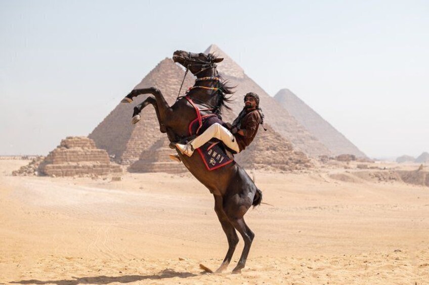 A person riding a horse in a desert with pyramids in the background. 1