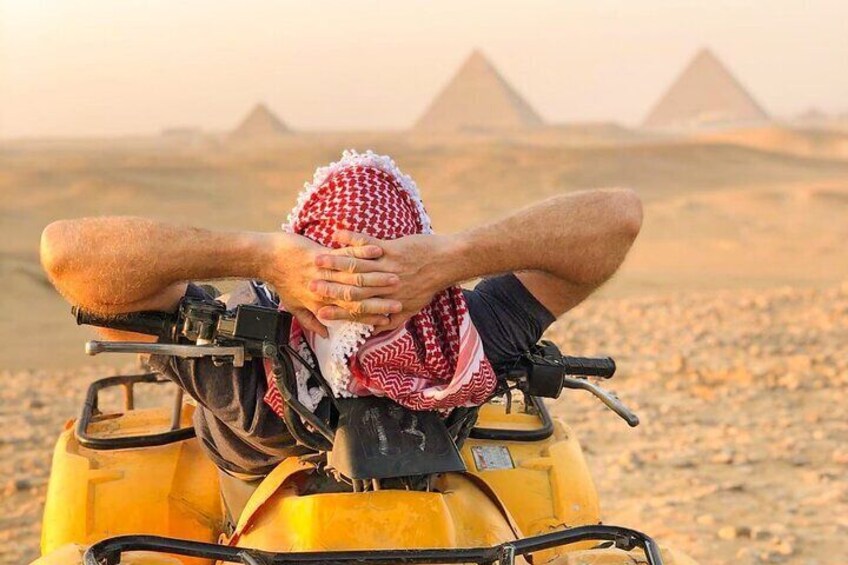A person on a yellow off-road vehicle in a desert with pyramids in the background. 3