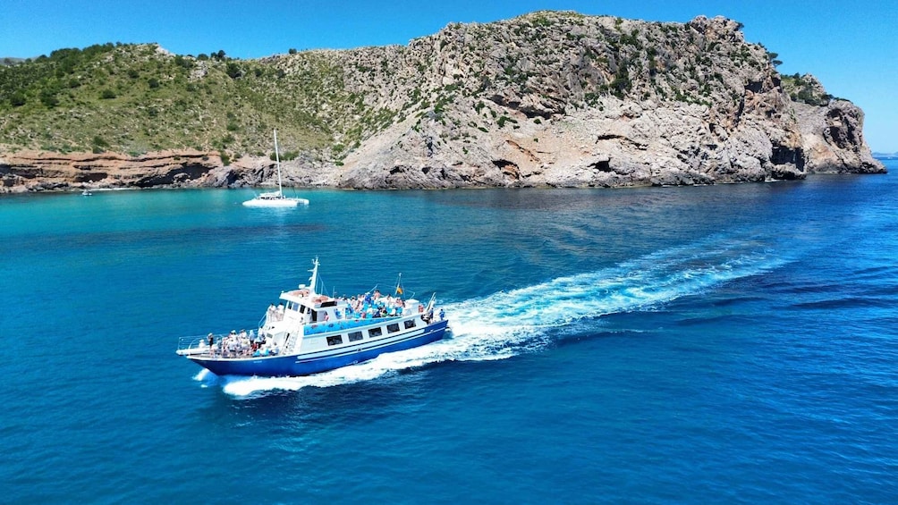 A blue boat with passengers on a clear day, moving through turquoise waters with a rocky coastline and a sailboat in the background. 1