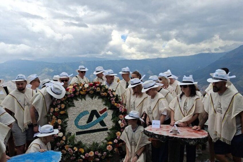 A group of people in white attire holding a floral wreath with a logo, standing on a mountain with a cloudy sky. 1