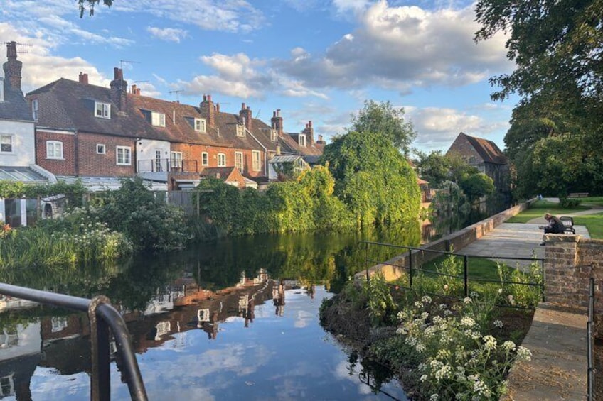 A calm river reflecting houses and trees, a clear sky, and a person sitting on a bench. 4