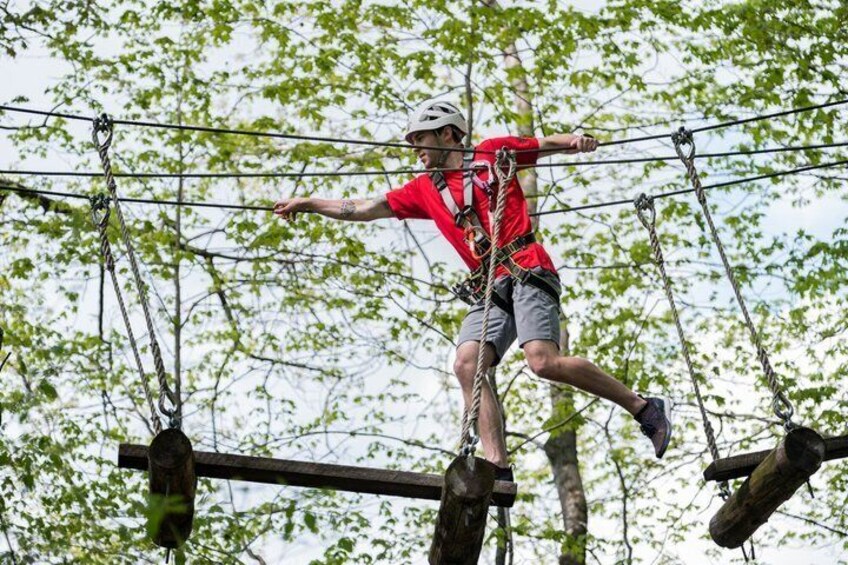 A person in a red shirt and helmet navigating a rope course with suspended platforms and safety harnesses. 4