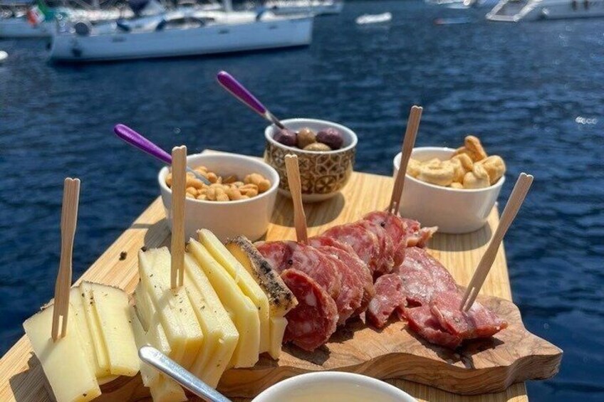 A wooden board with cheese, cured meats, and nuts by a body of water with boats in the background. 1
